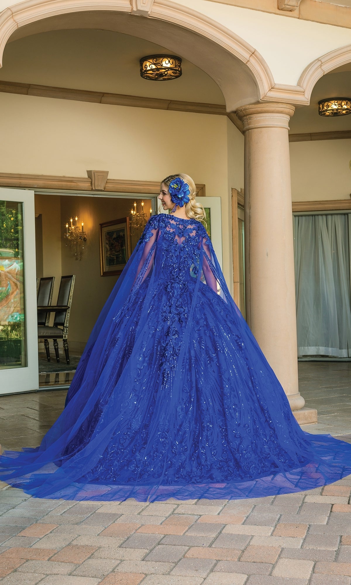 A woman stands outdoors before an ornate building, wearing Quinceanera Dress 1649 By Dancing Queen—a royal blue ball gown with a sweetheart neckline, long train, and matching headpiece.